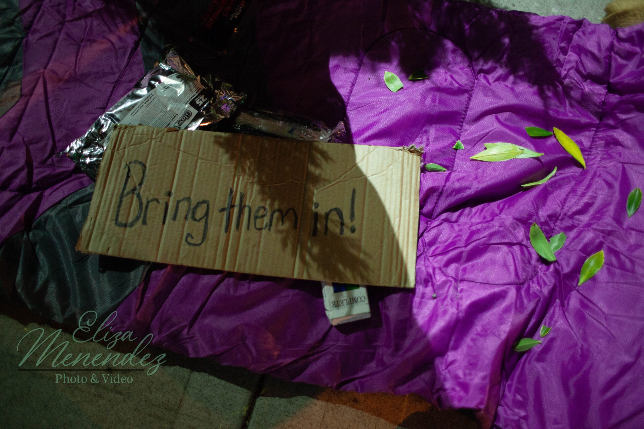 A sign that says, "bring them in!" on a sleeping bag on the sidewalk.